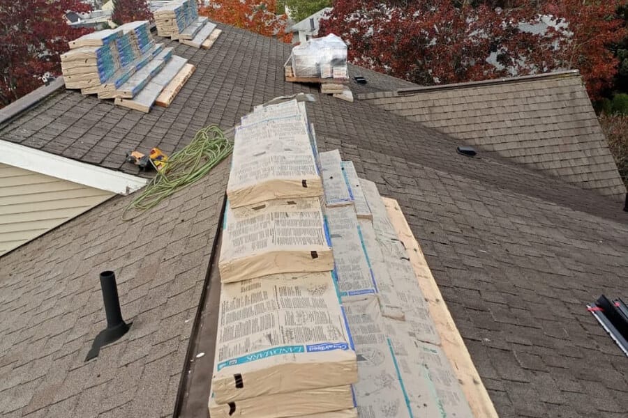 Bundles of shingles and rolls of underlayment stacked along the slope of a roof due to be replaced, with autumn trees behind.